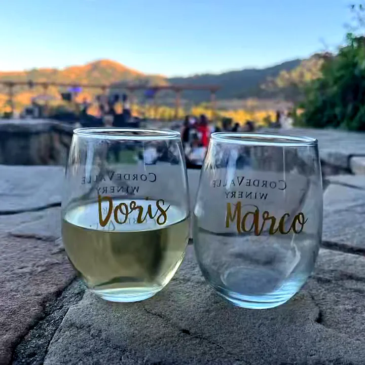Two stemless wine glasses personalized in gold with the names Doris and Marco, on a stone patio overlooking the hills at sunset